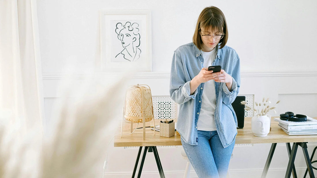 Woman in a denim shirt leaning against a wooden desk while looking at her smartphone in a bright, minimalist home office.